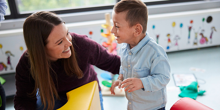 Betreuungssituation in einer Kita: Eine erwachsene Person begleitet ein spielendes Kind mit Behinderung in einem farbenfrohen Gruppenraum.