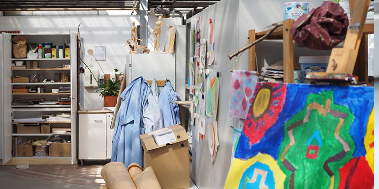 Atelierraum mit bunten Kunstwerken, Malutensilien, Kitteln und Regalen voller Materialien.