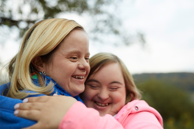 Zwei Menschen mit Behinderungen umarmen sich im Freien vor einer landschaftlichen Kulisse.