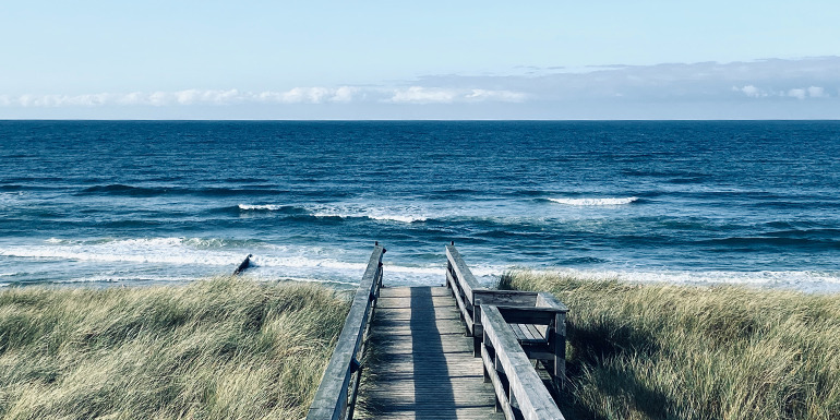 Holzsteg führt durch Dünen zum Meer mit Wellen und weitem Horizont.